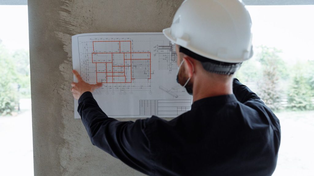 Mann mit Schutzhelm hält einen Bauplan an eine Wand