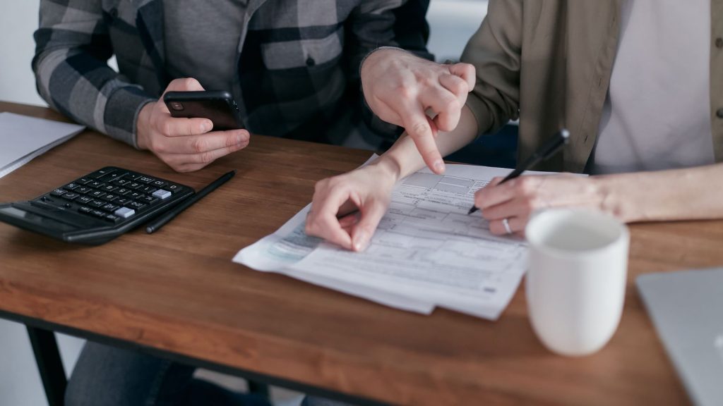Hände zeigen auf ein Formular, daneben Taschenrechner, Smartphone und Stift auf einem Holztisch